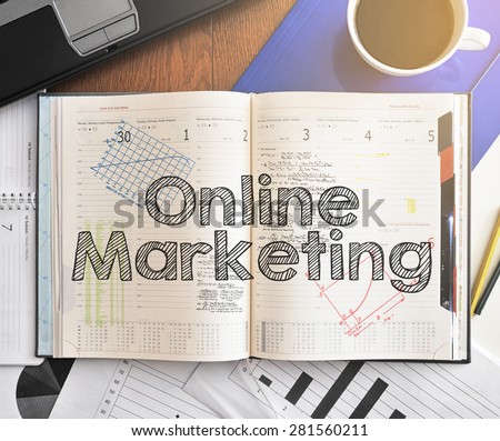 Notebook with text inside Online Marketing on table with coffee, laptop and some sheet of papers with charts and diagrams