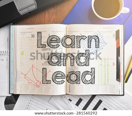 Notebook with text inside Learn and Lead on table with coffee, laptop and some sheet of papers with charts and diagrams