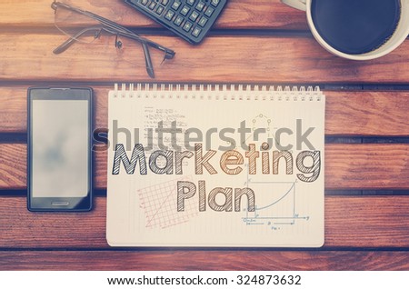 Notebook with text inside Marketing Plan on table with coffee, mobile phone and glasses.