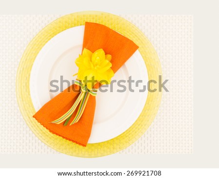 Bird\'s eye view of simple place setting arrangement with yellow daffodil flower, orange napkin, white plate and yellow charger on woven placemat.