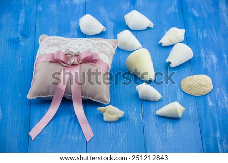 Beige wedding rings pillow with pink ribbon and shells on wooden vintage blue background