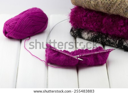 Knitting needles with a ball of woolen threads and colorful knitted woolen sweaters on white wooden background