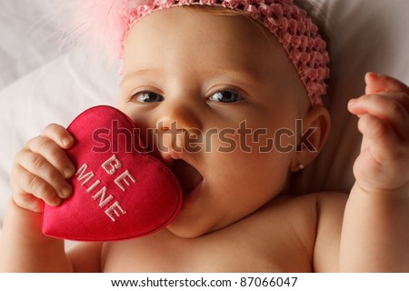 Valentine\'s day picture of baby chewing on a heart that says Be Mine on white fabric surface