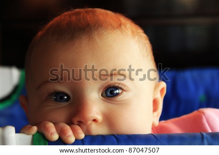Blue-eyed baby peeking over blue pack and play crib