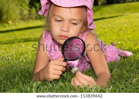 Little girl inspects looks at a flower through a magnifier while lying on a green lawn in summer park