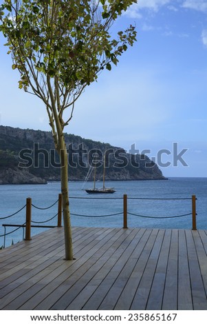 SANT ELM, MAJORCA, SPAIN - OCTOBER 30 2013: Seaside wooden deck with a tree and ocean view, a brig sailboat with two masts at sea on October 30 2013 in Sant Elm, Majorca, Balearic islands, Spain.