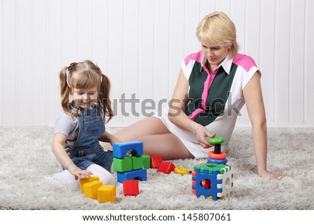 Happy little daughter and her pregnant mother play toys on carpet at home. Focus on woman.