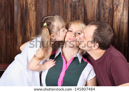 Father and little daughter kiss mother on bench next to wooden wall.