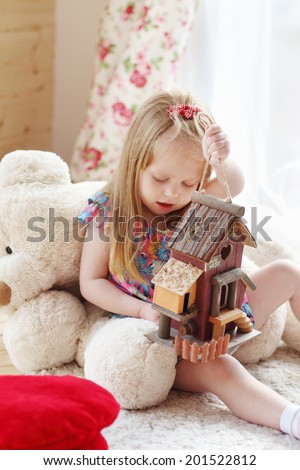 Pretty blonde little girl sits on carpet near window and holds toy house