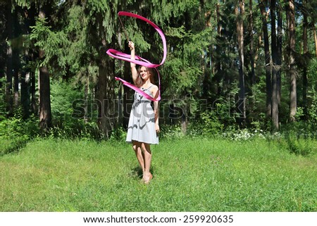 Beautiful girl in dress is engaged in rhythmic gymnastics with ribbon on grass on sunny day
