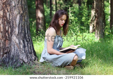 Beautiful young woman in dress sitting under tree on grass and reading book