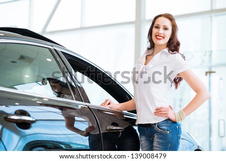 attractive young woman standing near a car in a showroom, chooses a car to buy