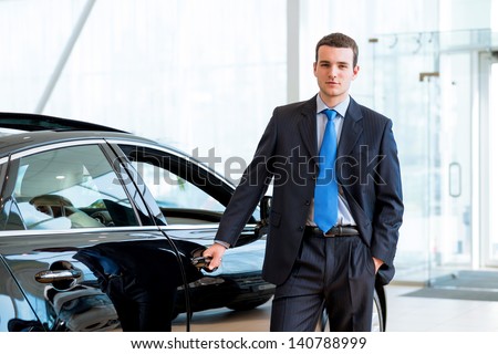 dealer stands near a new car in the showroom, put one hand on the car