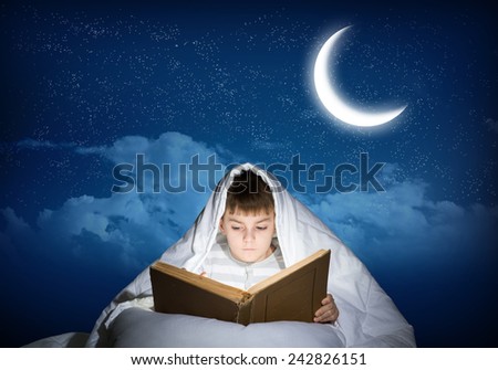 Cute boy in bed reading book under blanket