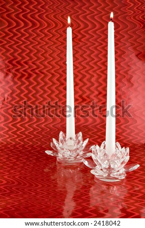 Two lit white taper candles in crystal candle holders on reflective red background