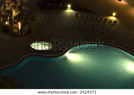 Partial view of blue swimming pool and hot tub at night with lights, palm trees and lounge chairs