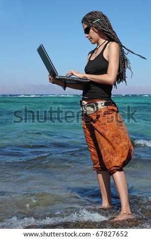 This young woman can be a professional freelancer or a student studying outdoors. She stands on a beach and work at her laptop.