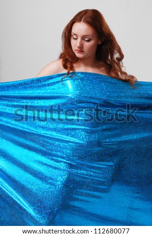 Nude woman is behind the knitted cloth. She is looking down. Female body is protruded through the piece of shiny material.
