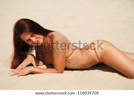 Young woman is lying on the sand. Her body is bent sexually. She is wearing tiny bikini. She is acquiring a tan in the sun.