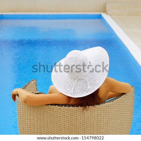Woman is sitting in the armchair near the outdoor swimming pool. She is photographed from back against the blue water background.