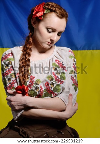 The sad young woman is wearing a shirt embroidered. Her red hair are plaited with a red ribbon. She is standing against the national Ukrainian flag.
