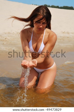 Pretty girl is sitting in the shallow water and drawing water. In the background there is the sand beach. She is wearing a white bikini. She has long hair and sexy body