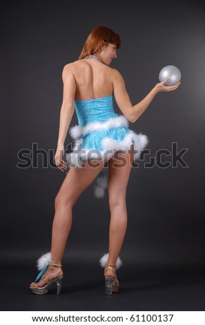 Young woman is stranding back. Pin-up is holding a silver christmas ball in outstretched arm on the dark background. She is wearing blue sexy suit decorated with white fluff.