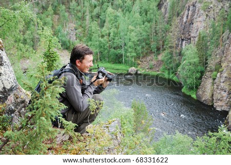 Happy photographer is sitting on the top of hill against the taiga forest and river full of rapids. The pleased tourist is holding a camera and smiling.