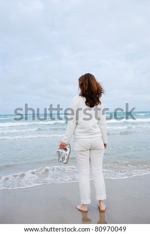 Barefooted woman is standing on the wet sand beach. She is wearing a windcheater and jeans. She is facing the blue sea and the cloudy sky. Waves are rolling in the flat seashore.