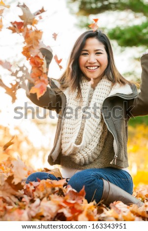 Young Asian woman in mid 20s playing in a pile of leaves at the park on an autumn day