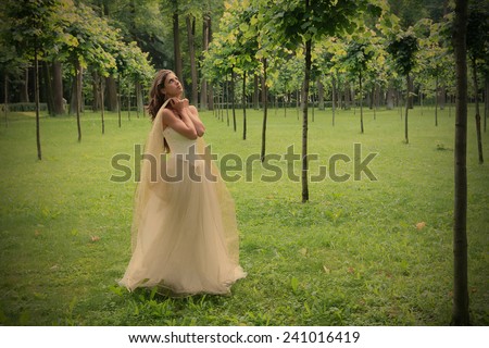 girl in white-golden gown in the summer park between young trees peers into sky, instagram image style