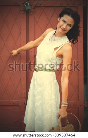 Smiling pretty middle-aged woman in a white dress with a bag standing near the old wrought-iron gate. instagram image filter retro style