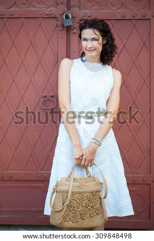 Smiling beautiful middle-aged woman in a white dress with a bag standing near the old wrought-iron gate