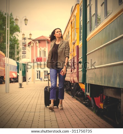 beautiful middle-aged woman with a suitcase on wheels and a passport in their hands. walking on railway platform along the passenger train. instagram image filter retro style