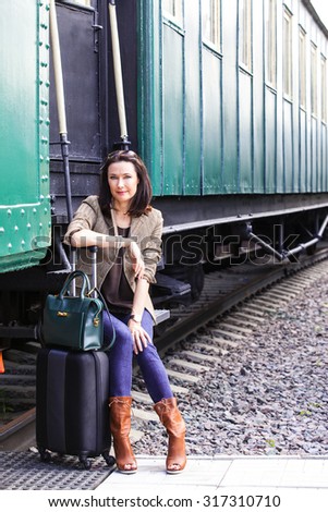 beautiful middle-aged woman with a suitcase and handbag sitting on the steps of the passenger railroad retro wagon waiting for the conductor. beginning of retro travel