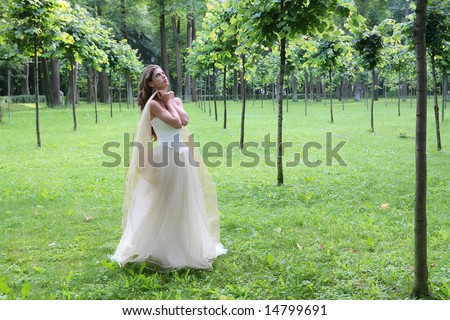 girl in white-golden gown in the summer park between young trees peers into sky