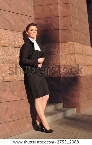 Beautiful businesswoman portrait standing outdoor full body and looking at camera with happy face. She smiles and holding black pad.