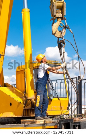 Construction worker in uniform and protective gear during hoisting works by mobile crane