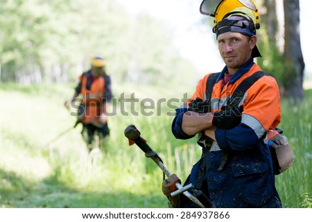 Portrait positive landscaper man worker with gas handheld string trimmer equipment during grass cutting team works