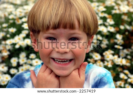 Smiling six year old boy being silly with hands under his chin outside in front of bush with white flowers.