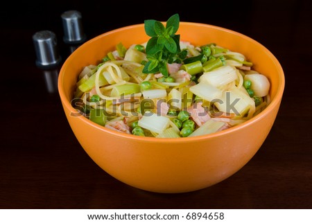 Healthy pasta dish with peas, leeks and bacon, in orange bowl on dark wooden table with salt and pepper shakers in background