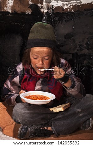 Poor beggar child eating charity food on the street sitting on cardboard plank