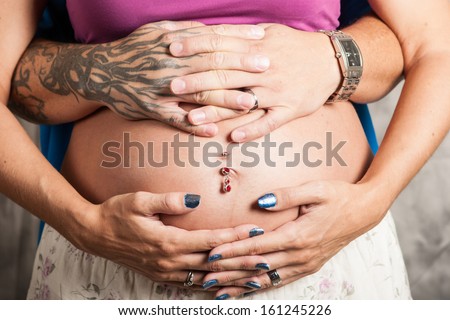 A pregnant woman\'s belly with pierced navel and holding hands with her tattooed spouse.