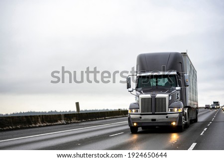 Big rig stylish industrial dark gray semi truck with turned on headlights transporting cargo in dry van semi trailer running on the twilight wet road with light reflection surface in rain weather 