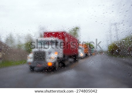 The convoy loaded big rig semi trucks with trailers and containers on the road in rainy weather through the windshield with rain drops.