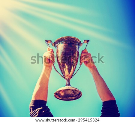 a man holding up a gold trophy cup as a winner in a competition toned with a retro vintage instagram filter effect app or action (backlit with the sun)