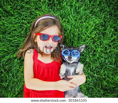 a cute little toddler girl with red sunglasses on holding a tiny chihuahua with blue goggles on laying in the grass in a park or backyard with a nice green lawn