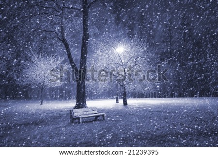 View of bench and shining lantern through snowing. Blue tone. Snow at night shot.