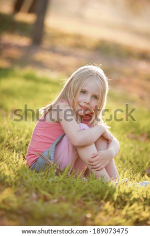 A young blonde girl sitting on the grass in the back light in summer.  She is sitting down smiling and looking at the camera.