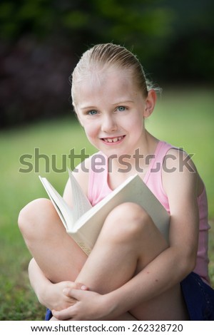 A young 7 year old girl relaxing with her white book outdoors.  She is relaxed studying or reading a novel.